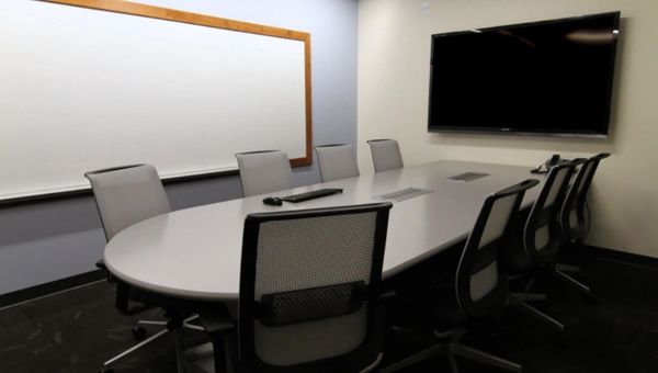 Modern conference room with oval table, mesh chairs, whiteboard, and large TV screen.