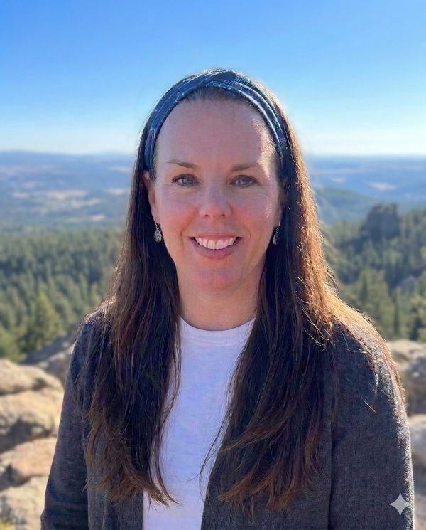 A smiling woman with long hair and a headband outdoors in a mountainous area.