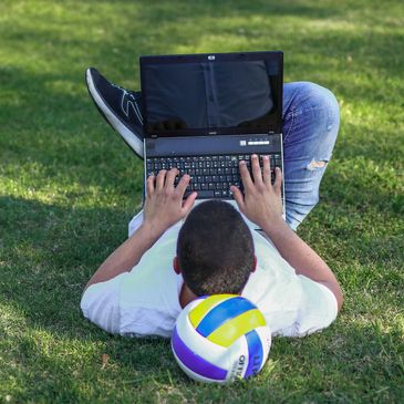 Person lying on grass using laptop with volleyball nearby.
