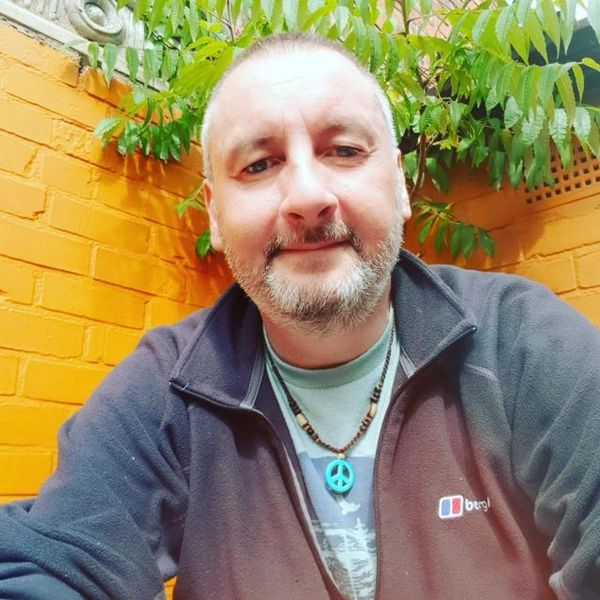 A man with a beard wearing a peace necklace and dark jacket in front of an orange brick wall.