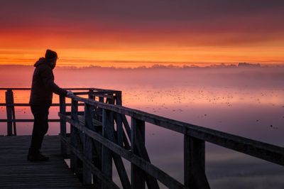 Person standing on a wooden dock at sunset over misty water.