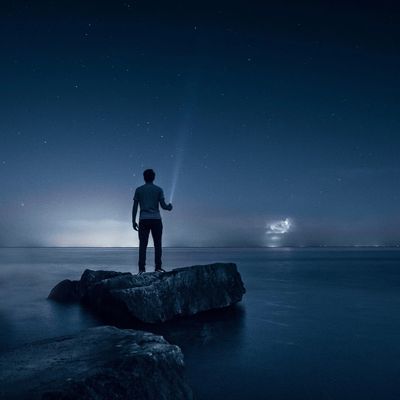 Man stands on a rock at night, shining a flashlight over calm water under a starry sky.