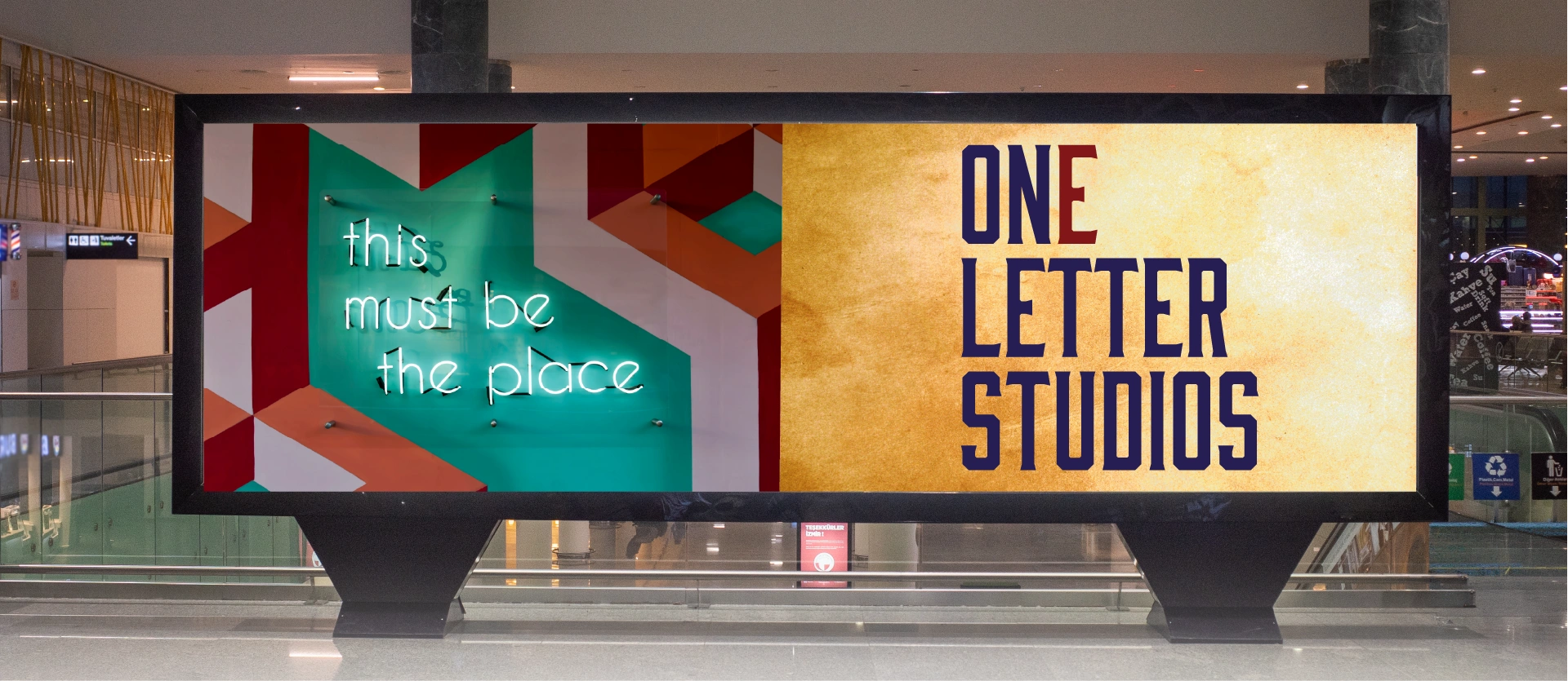 A large billboard in an indoor setting in a transit center. On the left of the billboard, there is a colorful neon sign that reads “this must be the place.” On the right, there is bold typography mostly in blue that reads, “One Letter Studios.” The “E” in the word “One” is set in red.