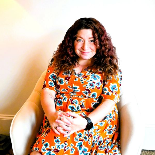 Smiling woman with curly hair in a bright floral dress sitting in a white chair.