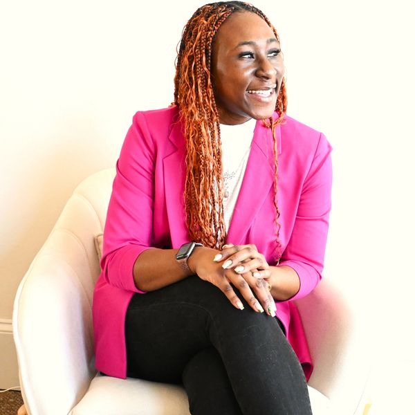 Smiling woman in pink blazer sitting on a white chair.