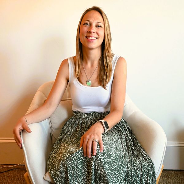 Smiling woman sitting comfortably in a white chair against a plain wall.