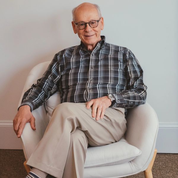 Elderly man in glasses sitting comfortably on a white chair, smiling gently.