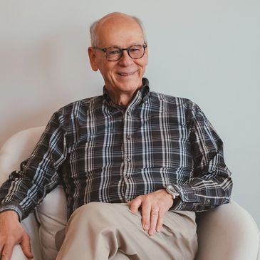 Smiling elderly man in glasses sitting comfortably in a white chair.