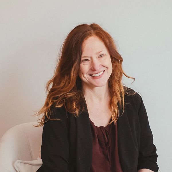 Smiling woman with long red hair sitting on a chair against a plain background.