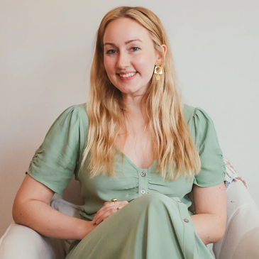 A smiling woman with long blonde hair wearing a green dress sitting on a white chair.