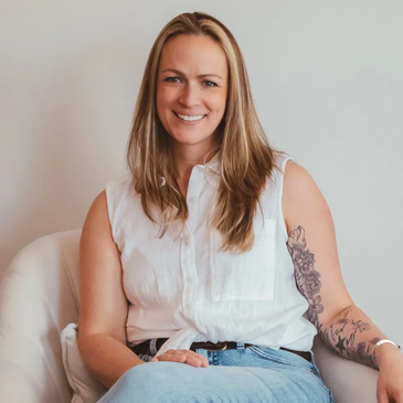 Smiling woman with long hair and a tattoo, seated comfortably in a white chair.