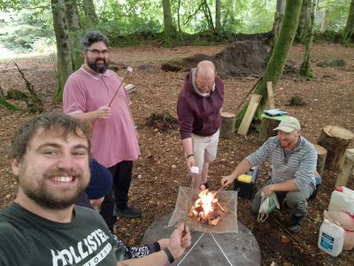 Four men enjoying roasting marshmallows around a small campfire in the woods.