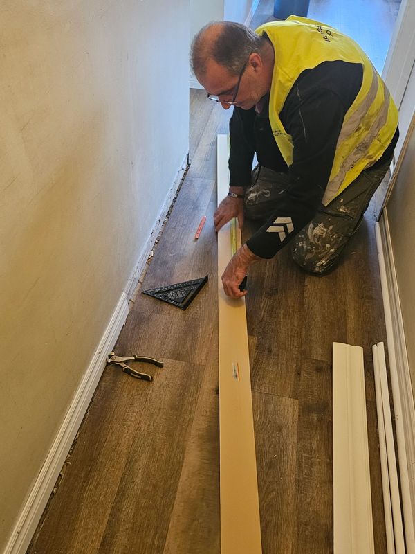 Man in a yellow vest measuring a wooden plank on the floor.
