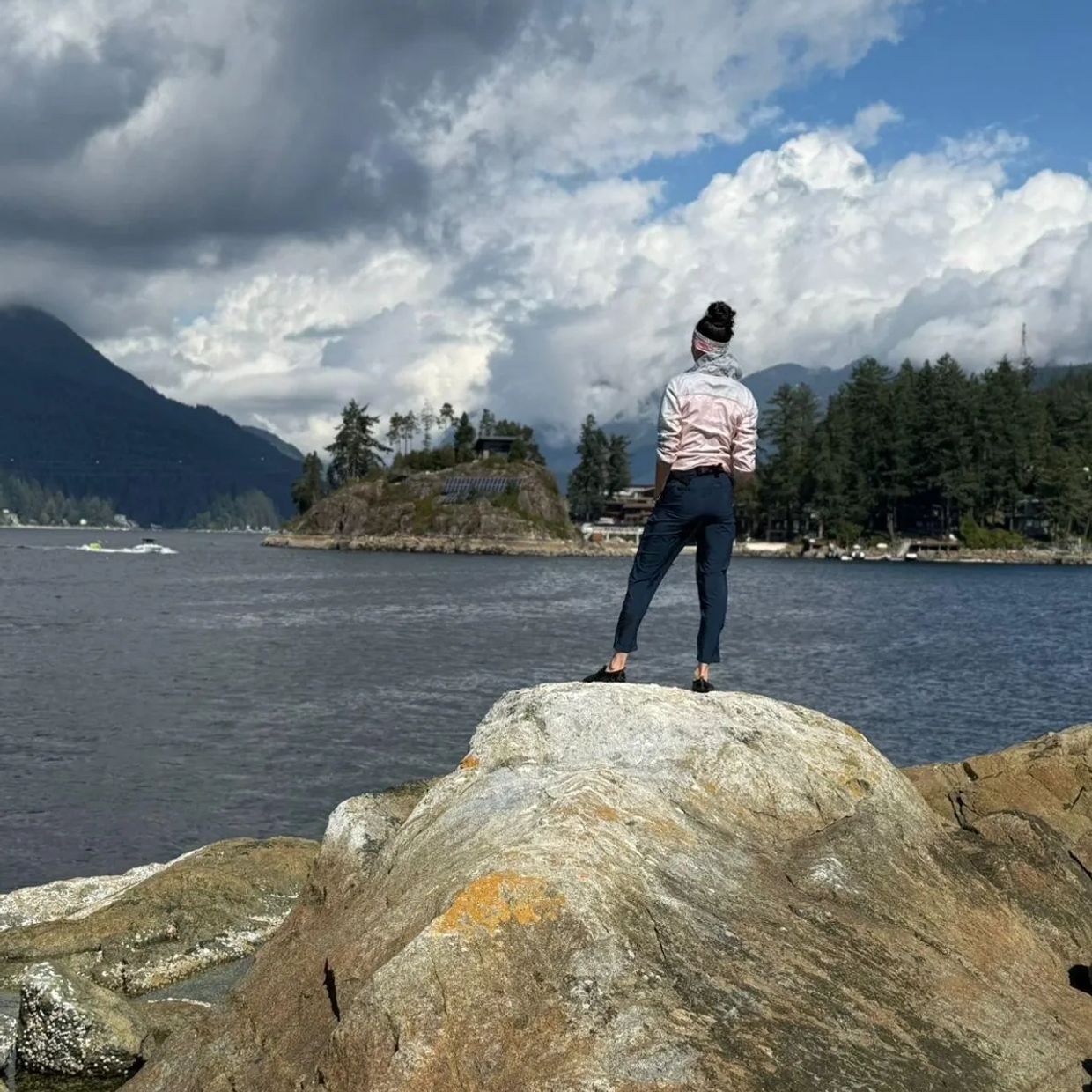Person standing on a rock overlooking a lake and forested mountains under a cloudy sky.