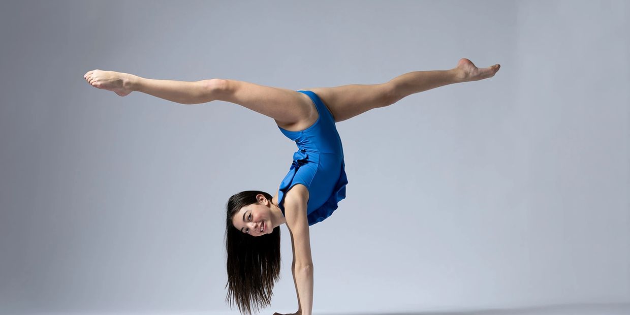 Young girl in blue leotard performing a handstand split against a plain background.