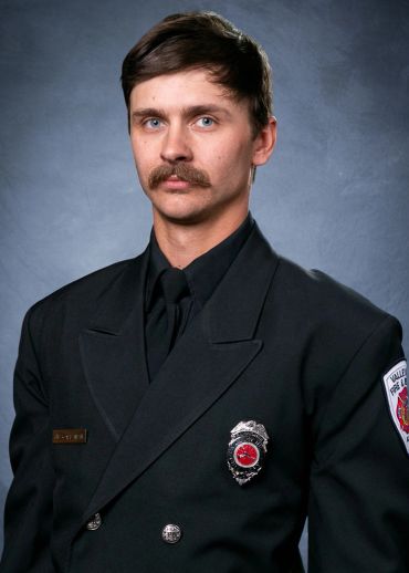 A firefighter in formal uniform with a mustache poses against a gray background.
