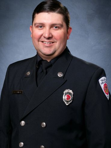 Portrait of a smiling firefighter in uniform against a blue-gray background.