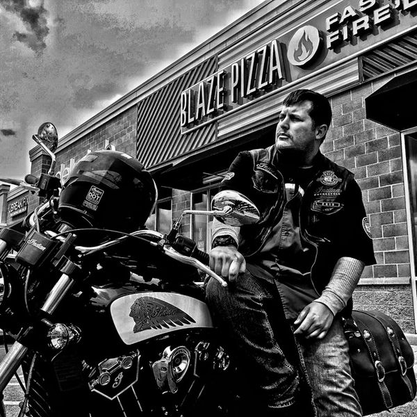 Man sitting on a motorcycle outside Blaze Pizza in black and white.