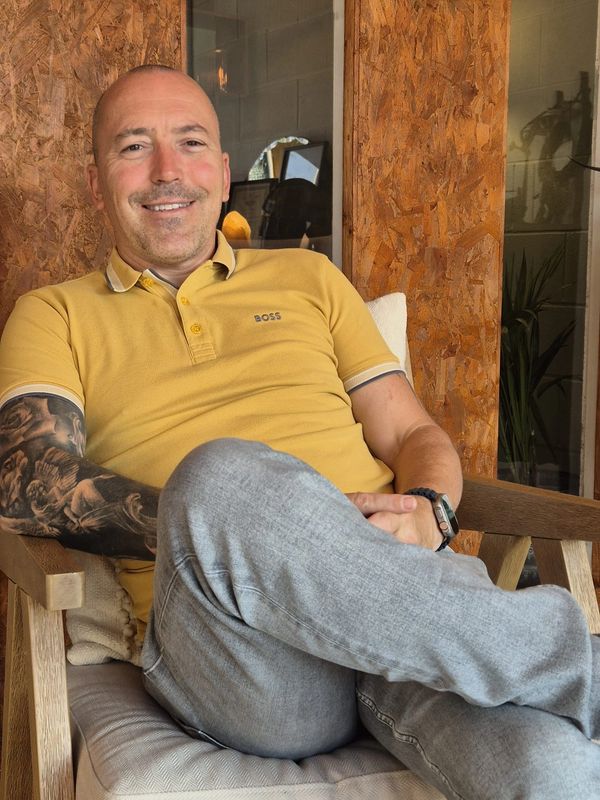 A man with a tattooed arm relaxes in a chair wearing a yellow polo shirt and gray pants.
