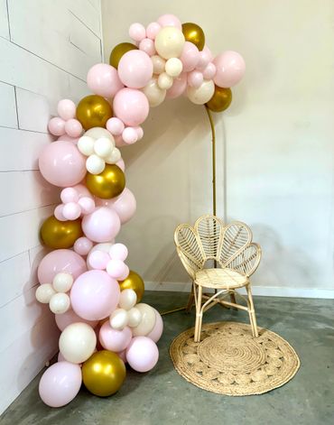 Elegant balloon arch in pink, white, and gold beside a wicker chair.