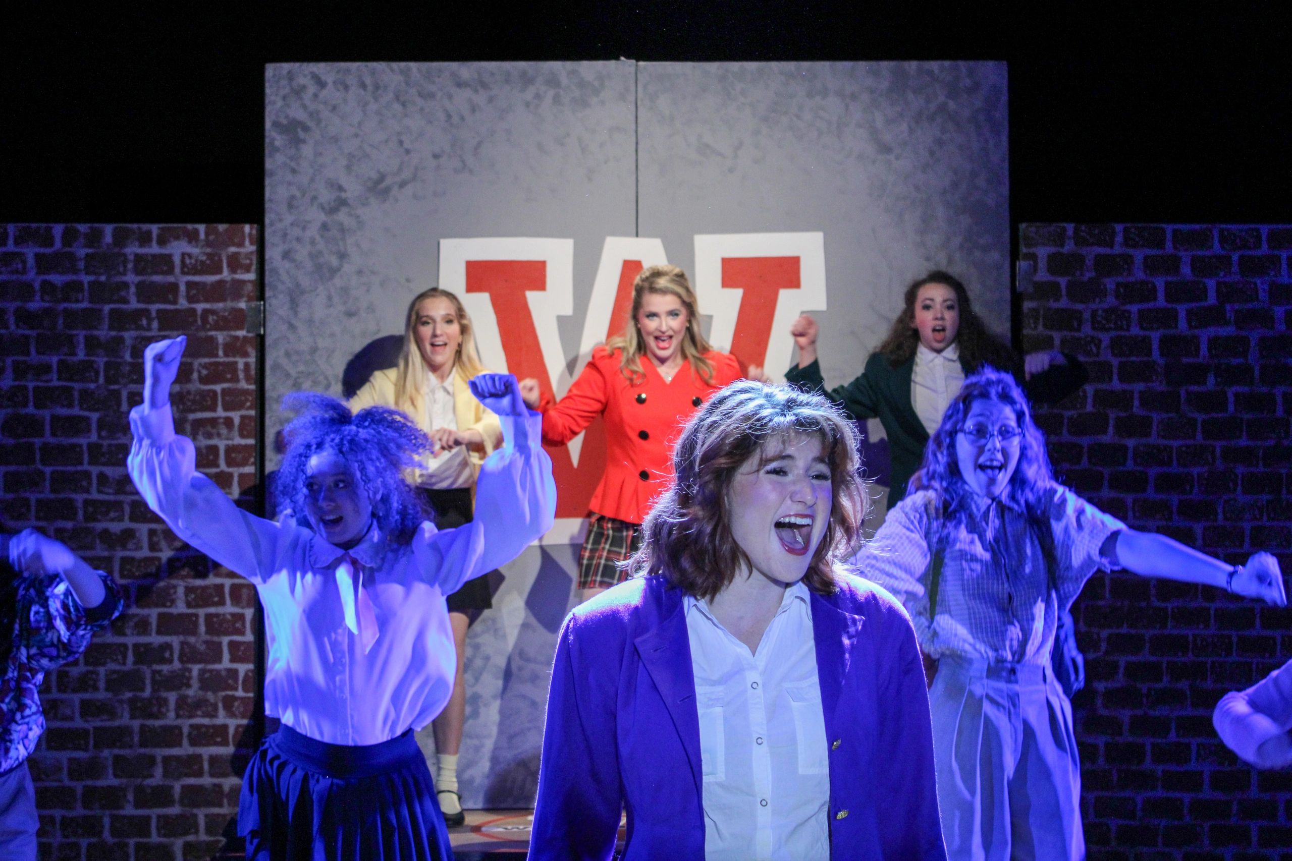 Actors perform energetically on stage with colorful lighting and bold costumes.