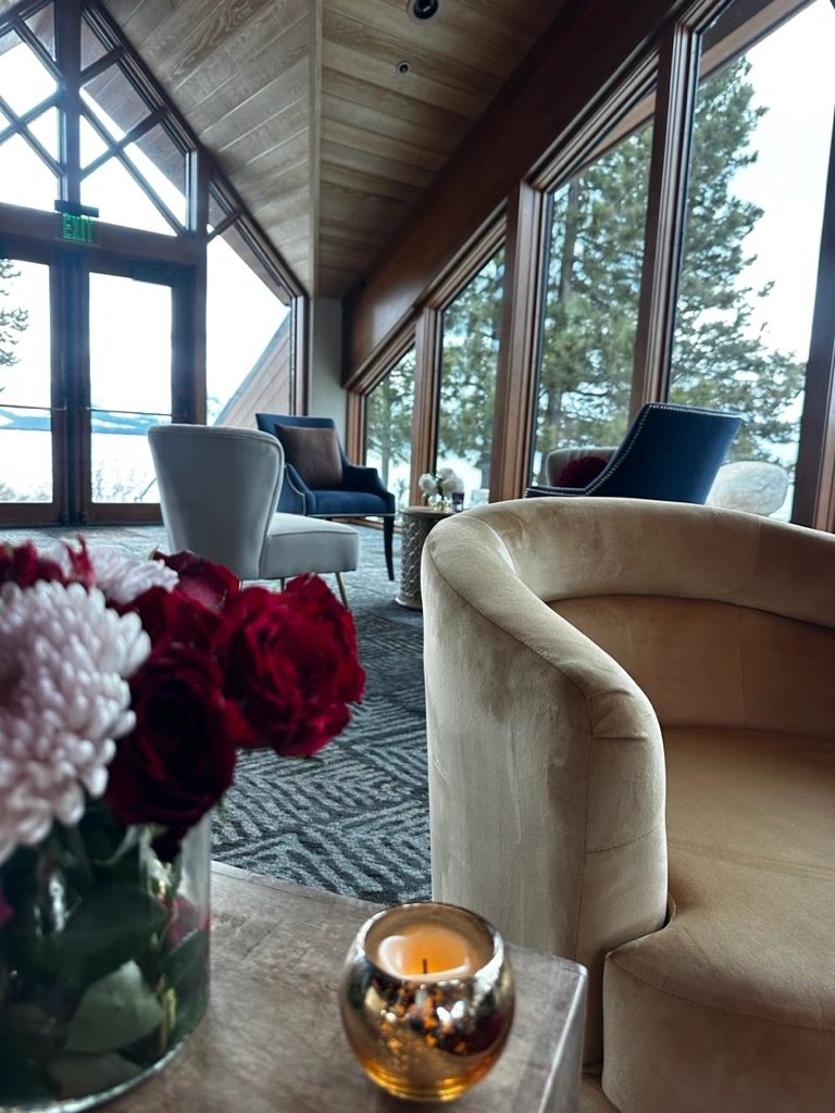 Lounge seating and flowers beside tall windows looking onto pines