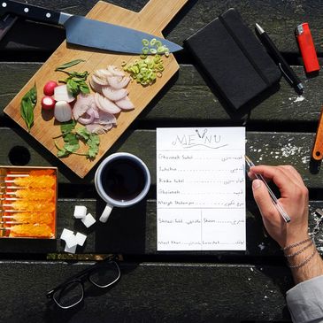 Person writing a menu next to sliced vegetables, coffee, and sugar cubes outdoors.