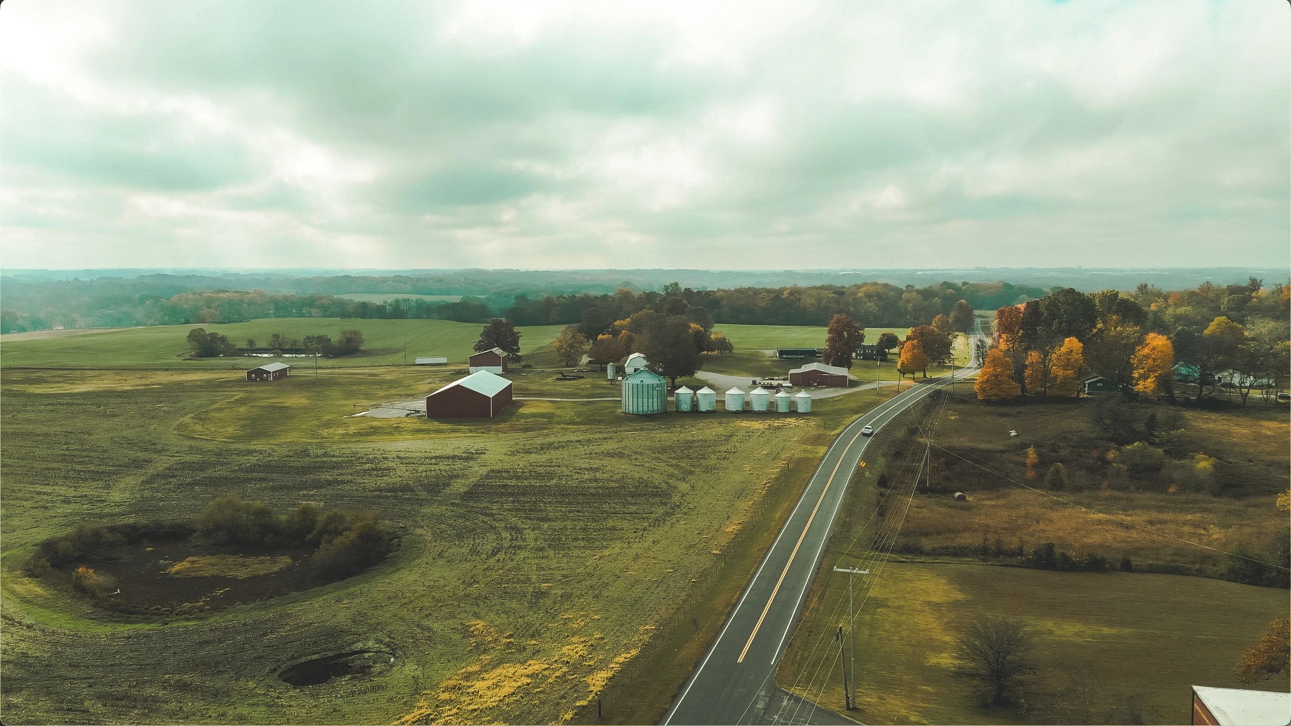 A rural road winds through farmland with silos and autumn trees.