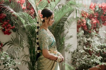 Woman in traditional attire with floral hair decoration stands beside lush greenery.