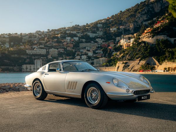 Classic silver sports car parked by the seaside with hillside town in the background.