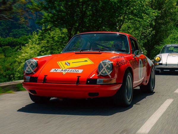 Classic red race car on a scenic road with a vintage white car behind.