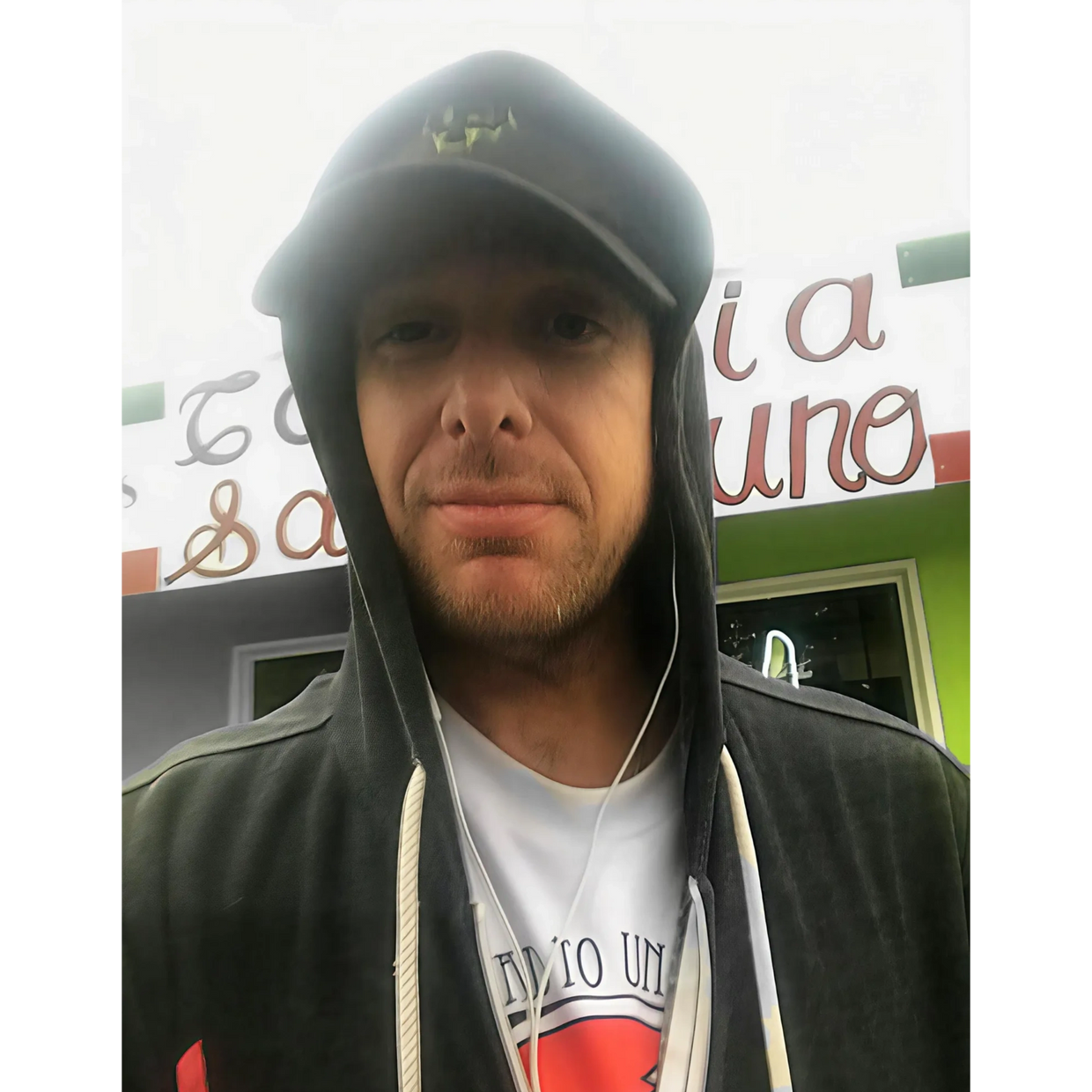 Man wearing a black hoodie and cap, taking a selfie outdoors.