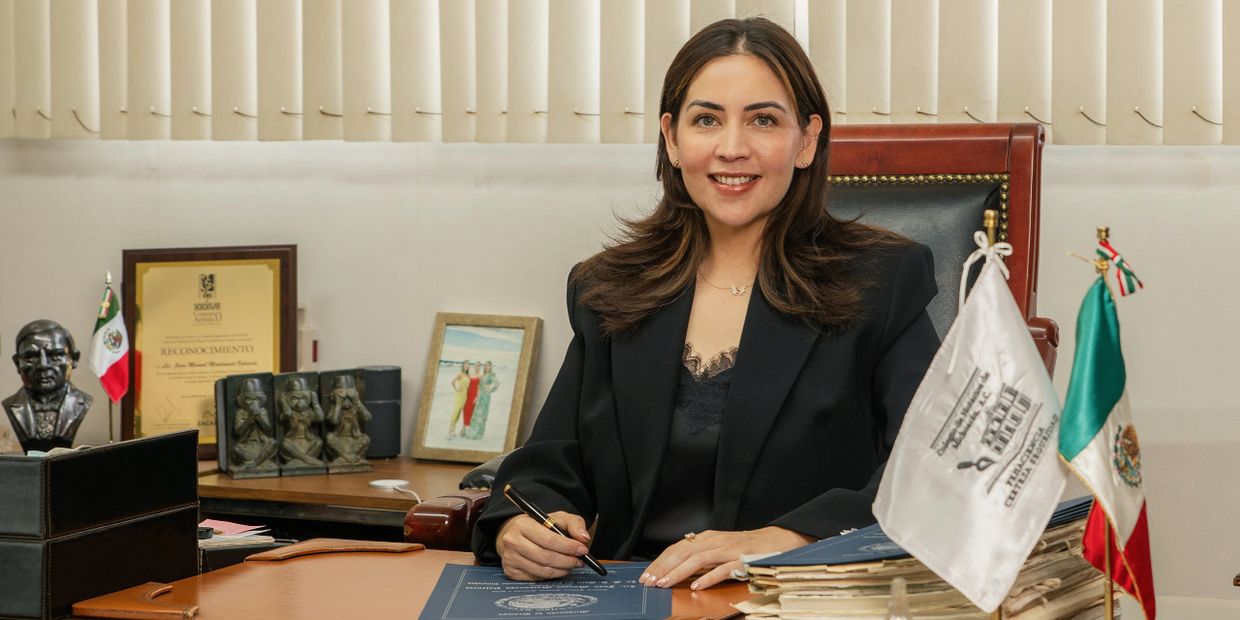 Woman in black blazer sitting at office desk, smiling and signing documents.