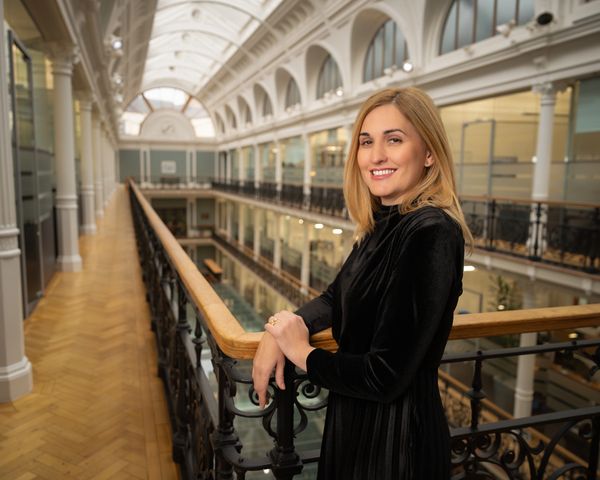 Smiling woman in black dress leaning on railing in a bright, elegant hallway.