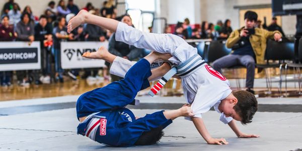 Two martial artists in gis competing on mats in a gymnasium.