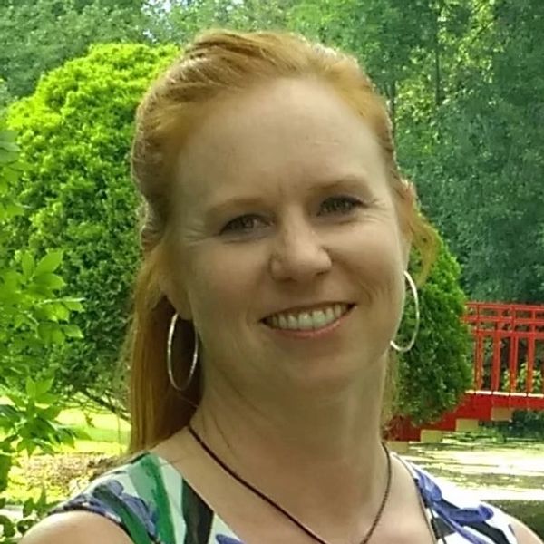 Smiling woman with red hair outdoors near green bushes and a red bridge.