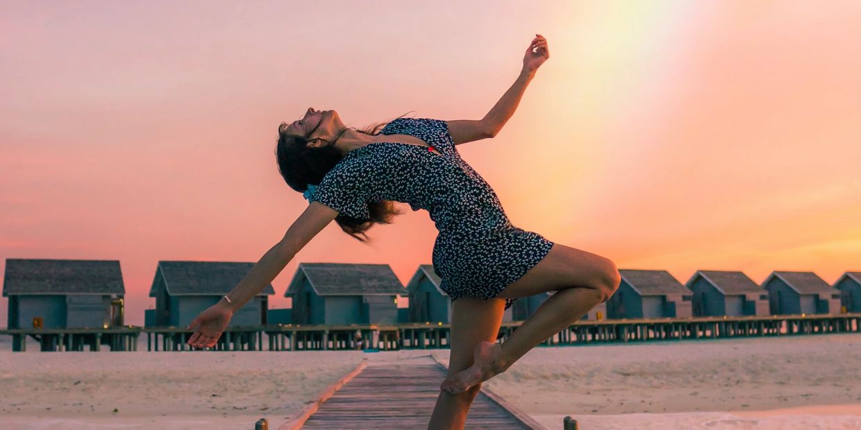 A woman dances joyfully on a boardwalk at sunset near beach huts.