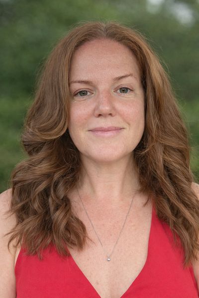 Woman with wavy auburn hair wearing a red top and a delicate necklace.