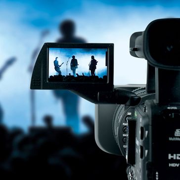 A video camera filming a live band performance on stage with blue lighting.