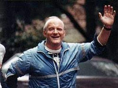 Elderly man in a blue jacket smiling and waving outdoors.