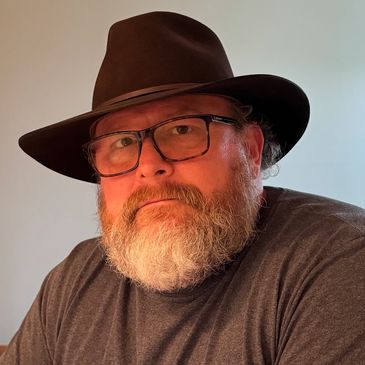 Bearded man wearing glasses and a dark hat, looking thoughtfully.