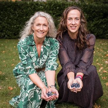 Two smiling women holding blueberries in their hands outdoors.