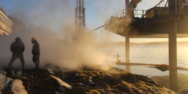 Two workers stand near a smoky coastal construction site at sunset.