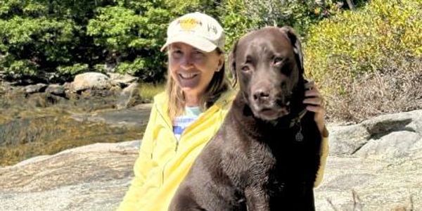 Woman in yellow jacket with a large chocolate Labrador on a rocky outdoor path.