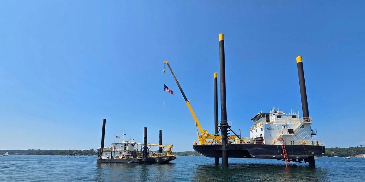 Two large industrial boats with tall black pillars and a crane, flying the American flag on a calm water body.