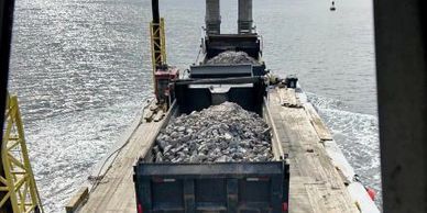 Dump trucks loaded with rocks on a barge at sea under a cloudy sky.