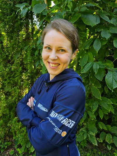 Smiling woman in a blue jacket stands with arms crossed in front of green foliage.