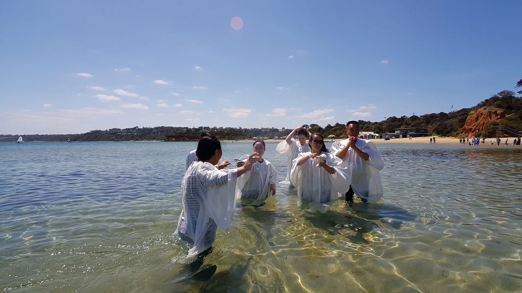 Group of people in white robes standing in clear shallow water at a beach.