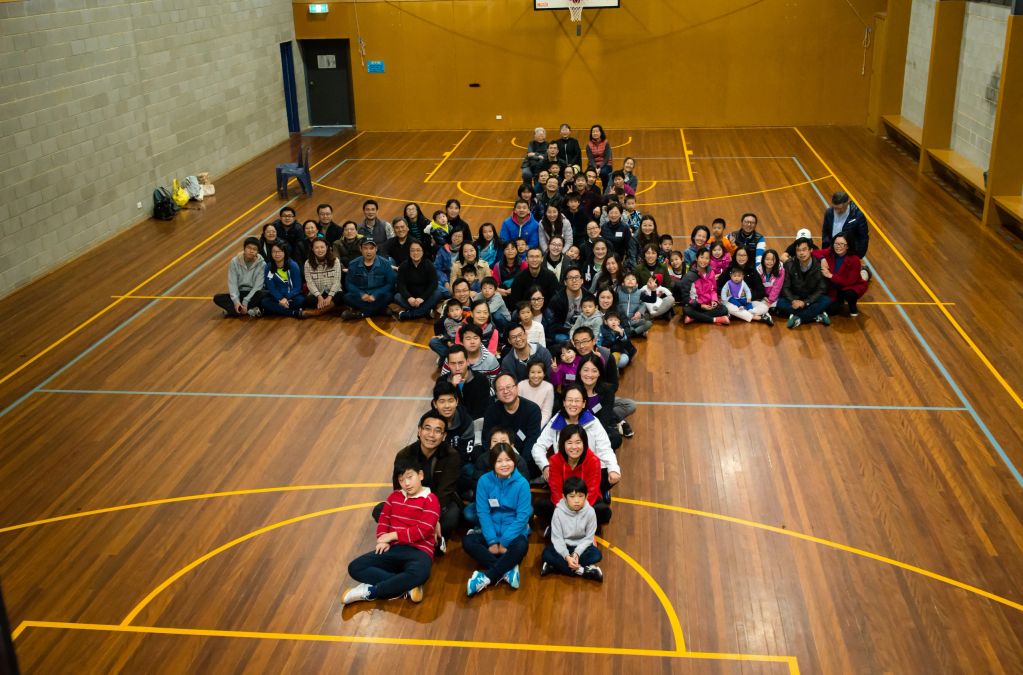 Large group of people sitting in a cross shape on a basketball court indoors.