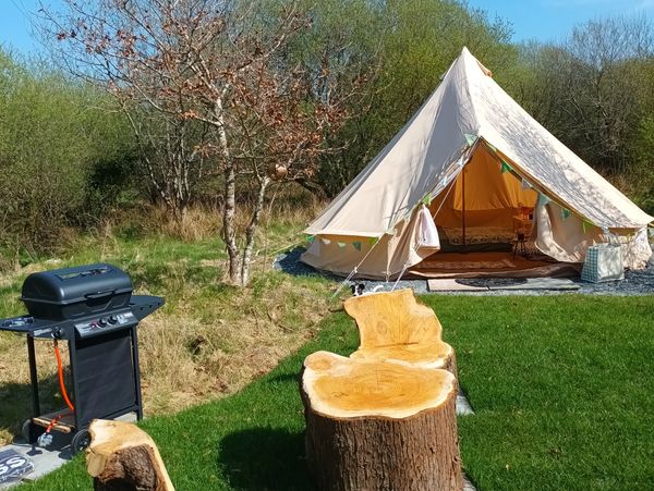 A cozy glamping tent with wooden stump seats and a barbecue grill outside.